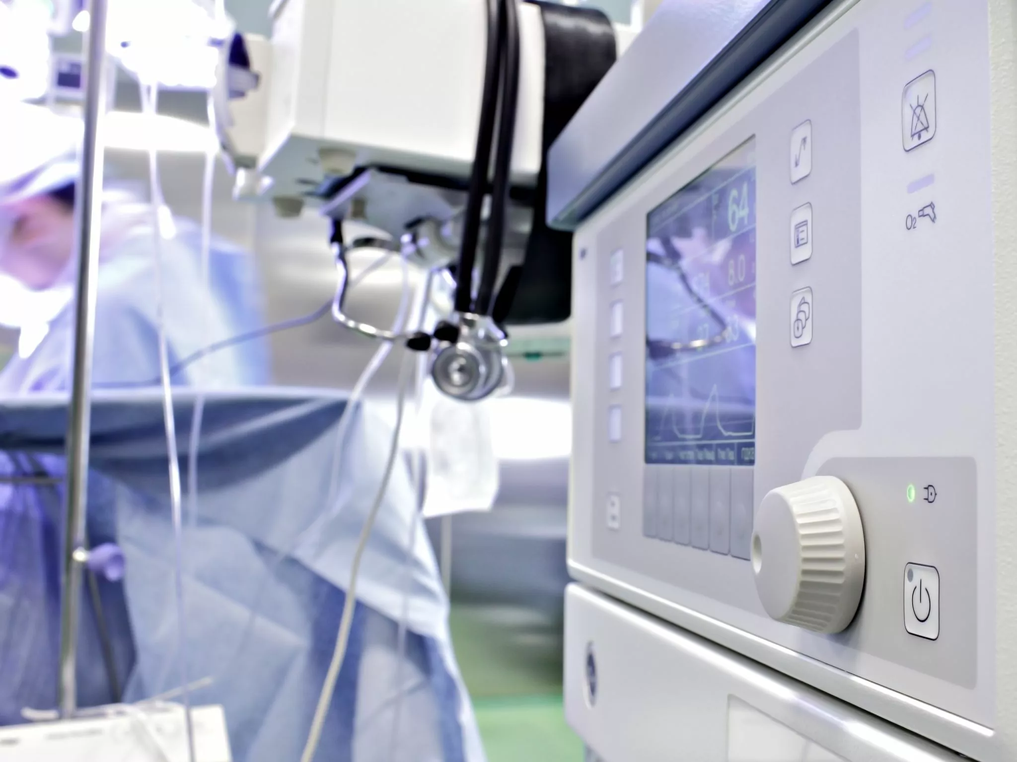 Close-up of an ISO 13485 certified medical monitor displaying vital signs in a hospital operating room, with blurry figures of medical staff in the background.